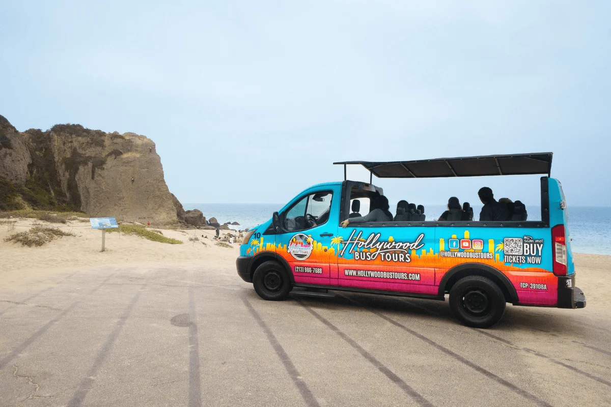 A colorful open-top Hollywood Bus Tours van parked in a lot beside tall coastal rock formations and sandy beach in Malibu.