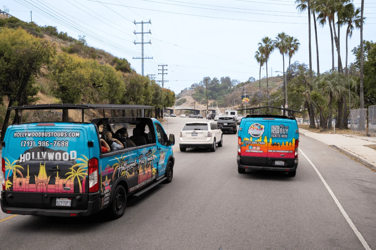 Two blue open-air tour vans filled with sightseers driving down a road lined with palm trees in the Hollywood Hills.