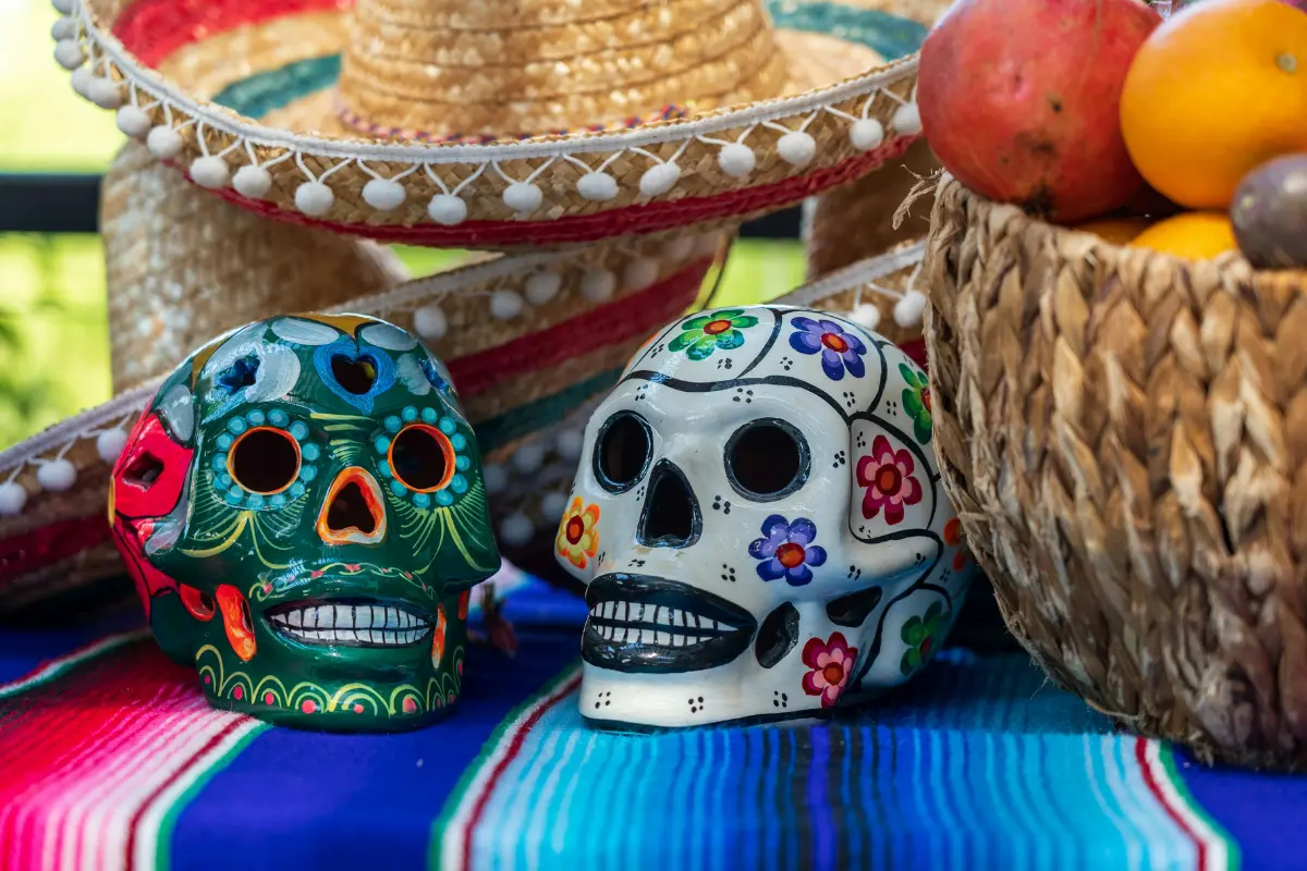 Two hand-painted ceramic sugar skulls with floral designs on a striped serape blanket in front of straw sombreros