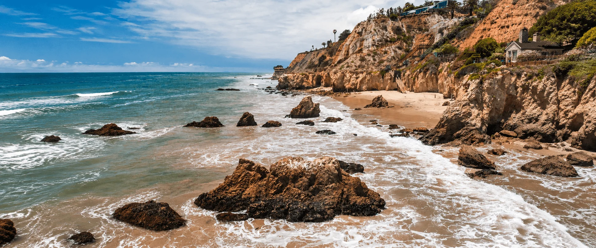 Panoramic view of ocean tides washing over rock outcrops in front of high coastal bluffs and luxury homes perched above El Matador State Beach