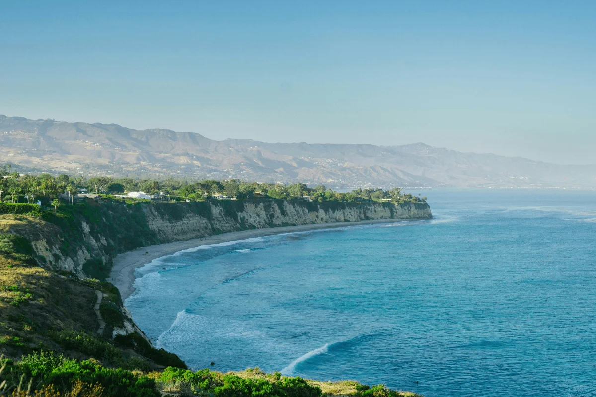 Elevated view showing the curved coastline, dramatic sea cliffs, and blue waters of Malibu stretching toward distant coastal mountains.