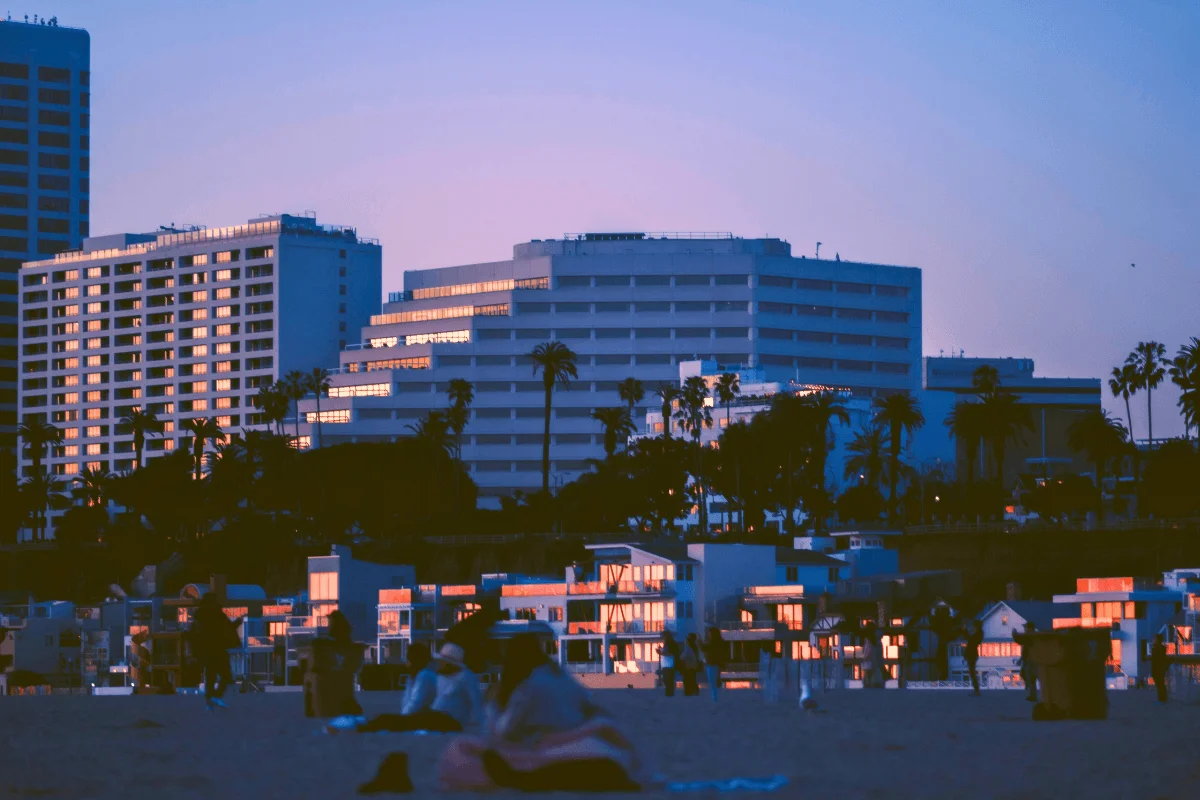 Beachgoers sitting on the sand looking toward illuminated high-rise hotels along the Santa Monica coastline during twilight.
