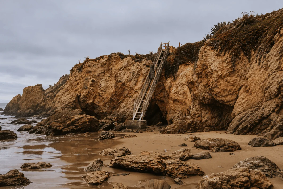 Steep wooden stairs built into towering cliff faces leading directly onto the rocky beach near natural sea cave openings.