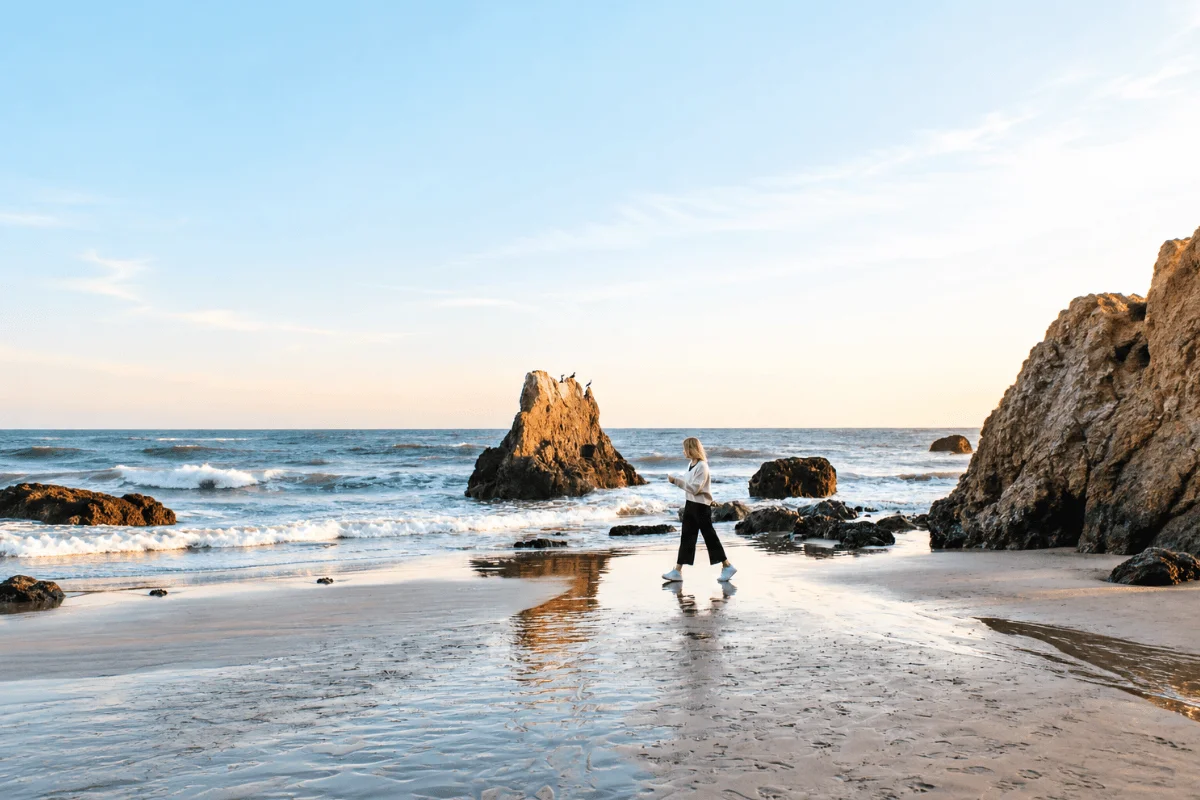 A person walking along the wet sand near large rock formations and sea stacks as gentle waves lap the shore at El Matador Beach.