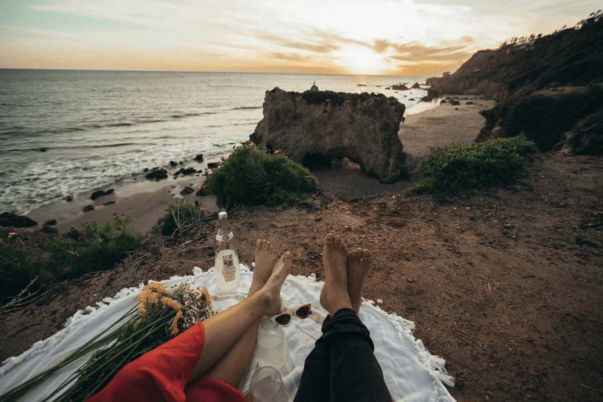 First-person view of a couple enjoying a blanket picnic on a bluff top overlooking a large sea arch rock formation at sunset.