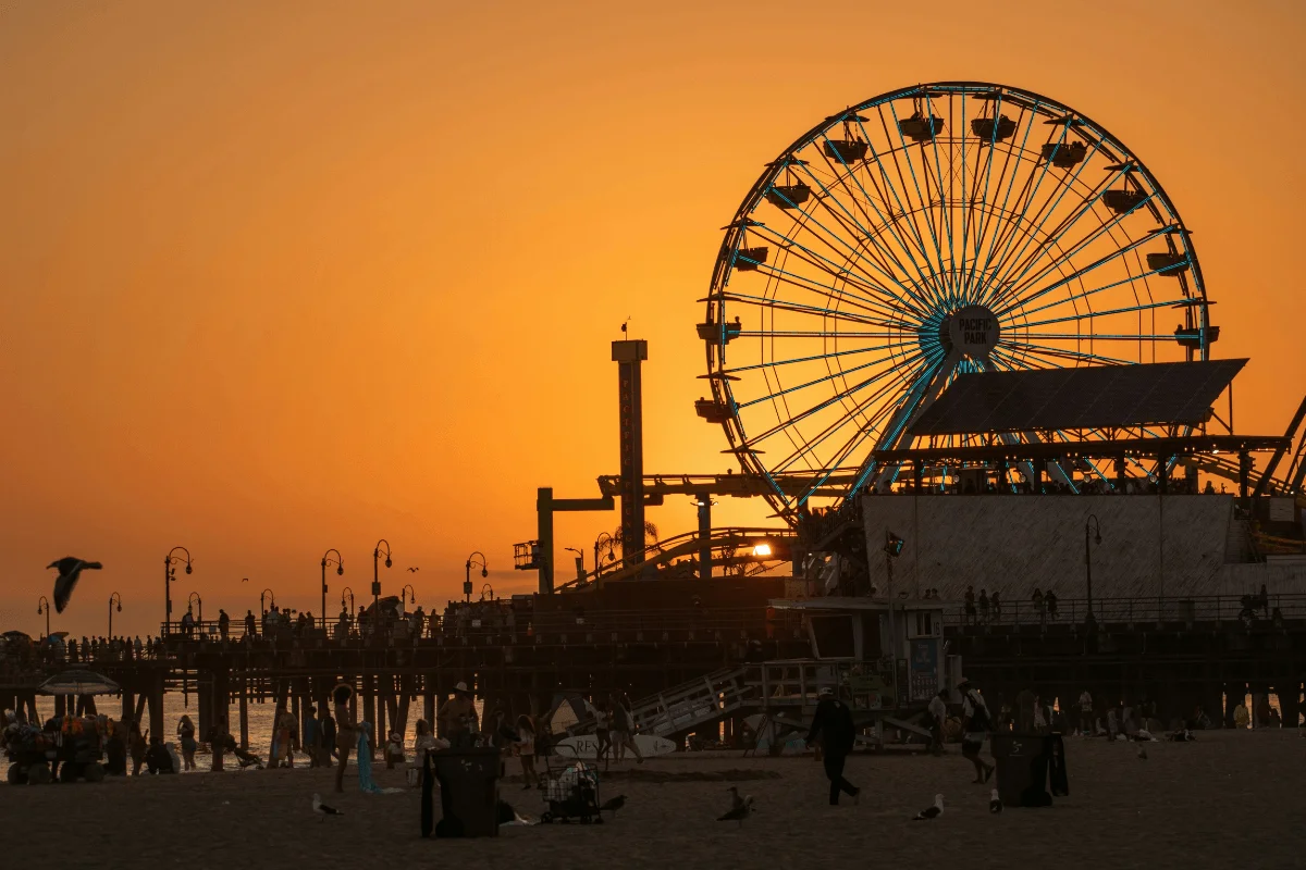 Silhouette of the Pacific Park Ferris wheel and roller coaster on Santa Monica Pier against a vibrant orange sunset sky.