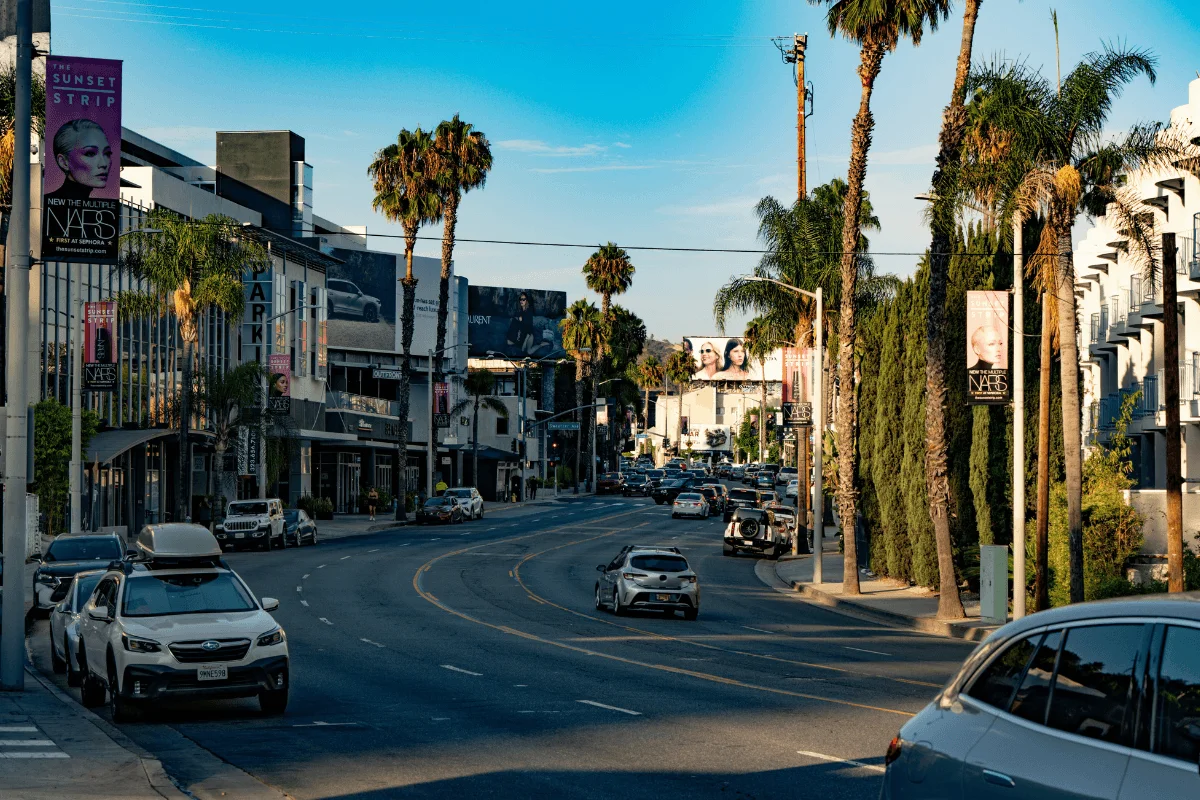 Cars driving down Sunset Boulevard lined with tall palm trees, modern buildings, and billboards under a clear blue sky.