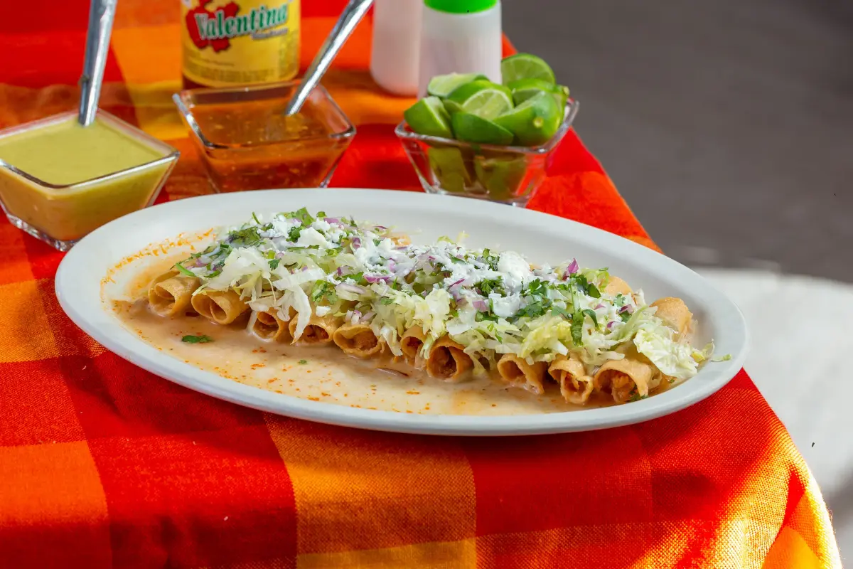 Plate of crispy taquitos topped with lettuce, cotija cheese, and red onion, served with green and red salsa and fresh lime wedges
