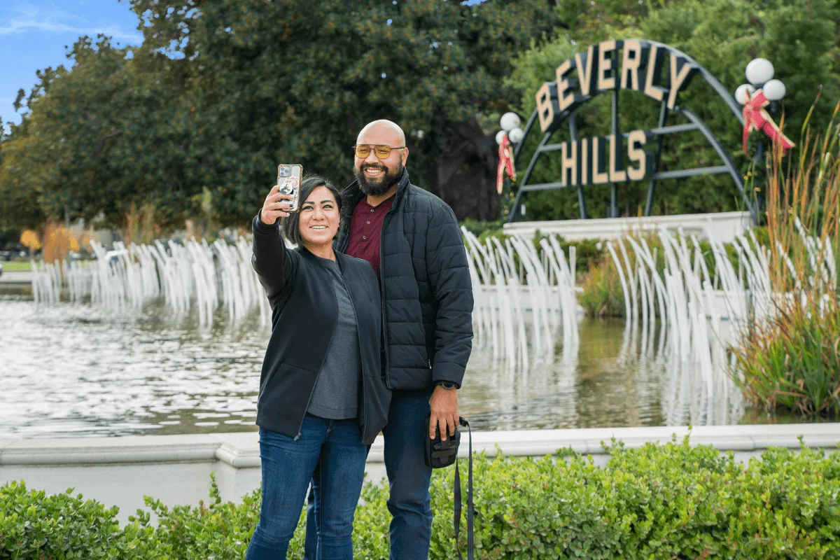 A smiling couple standing in front of the iconic arched Beverly Hills sign taking a selfie with a smartphone.